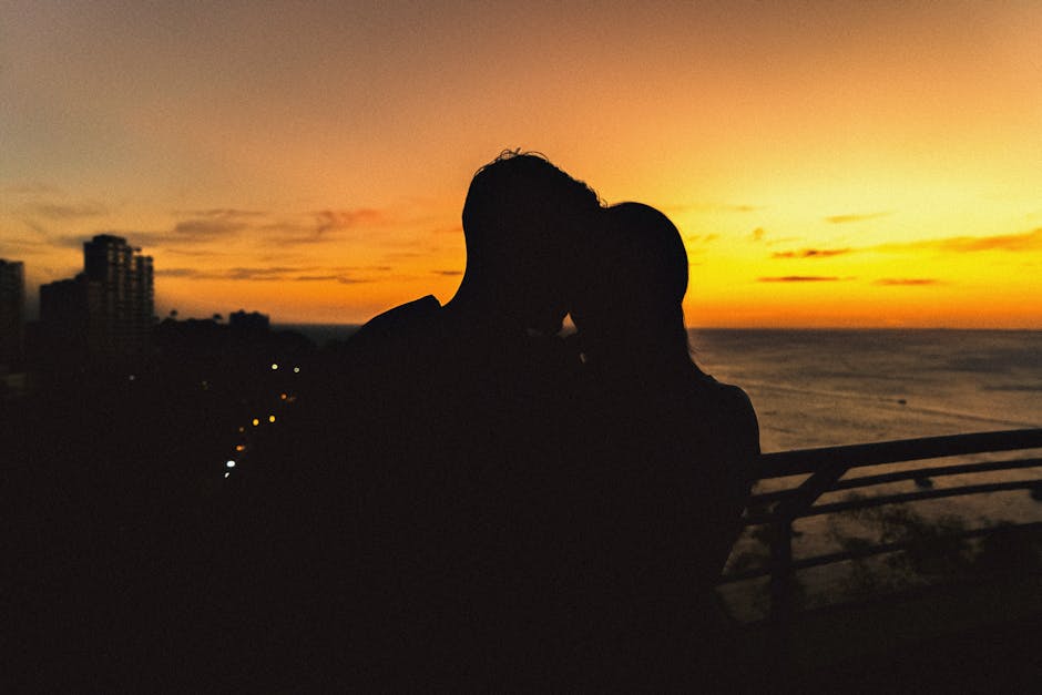 Silhouette of a couple sharing a romantic moment at sunset by the seaside with city lights in the background.