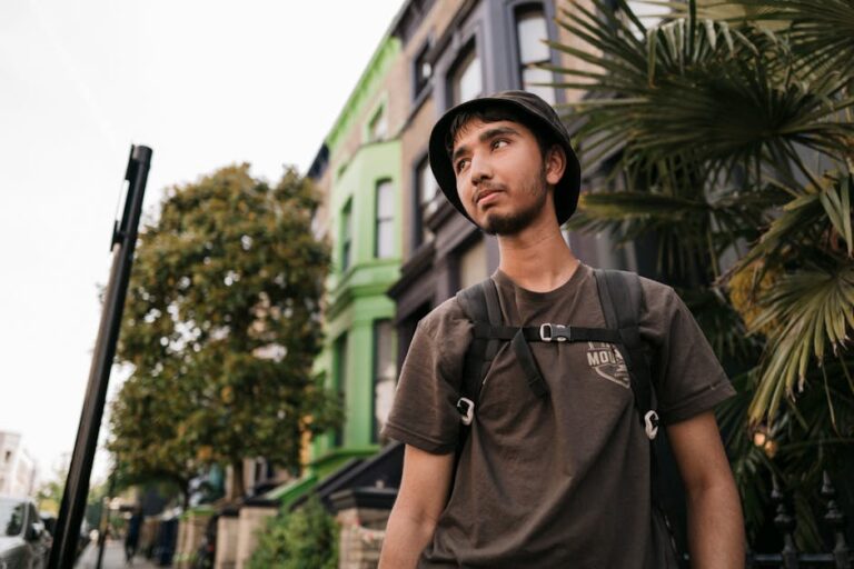A young man explores London, wearing a backpack and bucket hat, next to urban greenery.