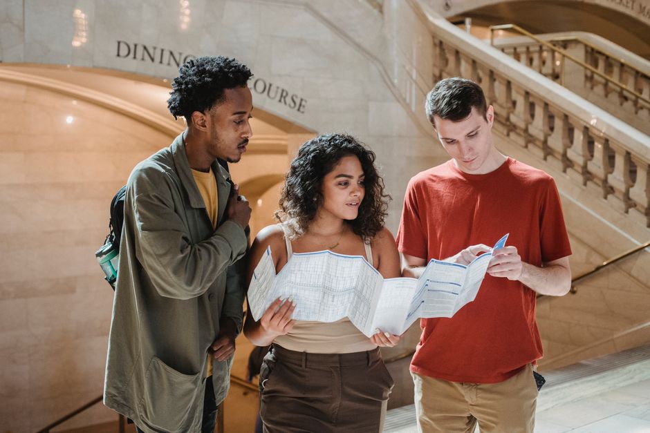 Focused young man pointing at map while searching for route with multiracial friends in Grand Central Terminal during trip in New York