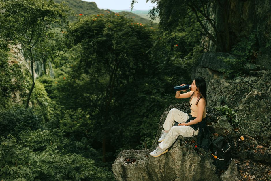 Woman sitting on a rock, drinking water in a lush green forest, symbolizing adventure and relaxation.