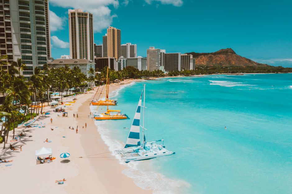 Aerial view of Waikiki Beach featuring a catamaran and Honolulu cityscape under a bright blue sky.