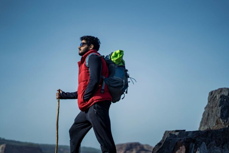 Man hiking in the wilderness with a red vest, trekking pole, and backpack against a clear sky.