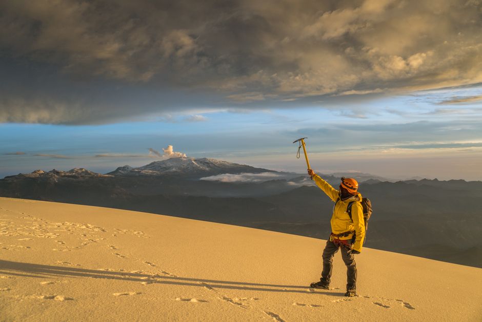 A climber triumphantly raises an ice axe atop a snowy peak with dramatic skies.